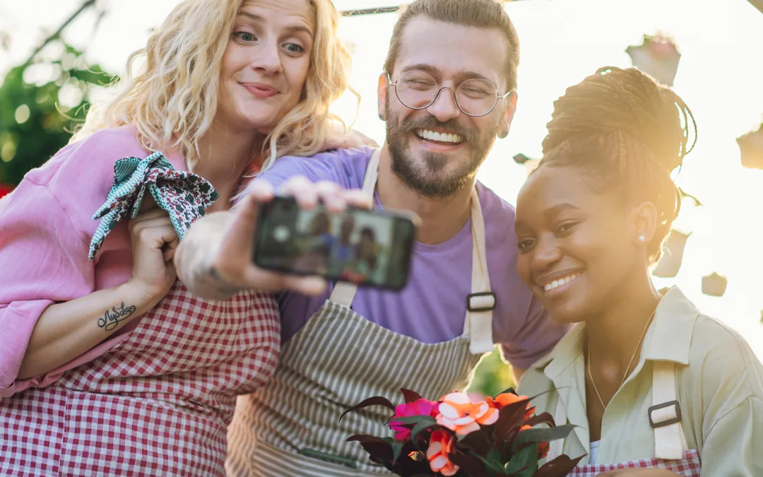 Group of happy multiracial people working with flowers at industrial greenhouse and taking a selfie with a smartphone. Lifestyle, gardening, profession and people concept. Diverse people. Copy space.